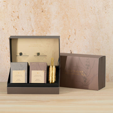 An open gift box with two boxes of tea inside next to a gold tea infuser. There is a closed brown box behind the open box.