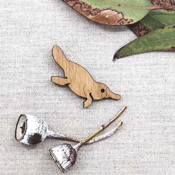 A wooden magnet in the shape of a platypus with gum leaves in the background.