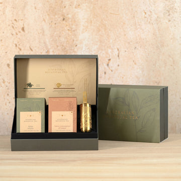 An open gift box with two boxes of tea and a gold tea infuser inside. There is a closed green box behind the open box.