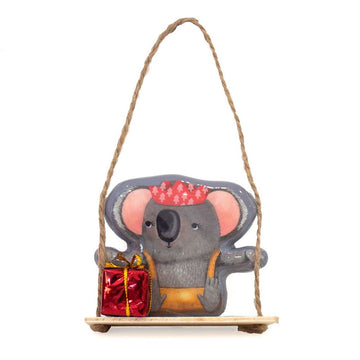 A Christmas decoration of a koala sitting on a swing holding a red present.