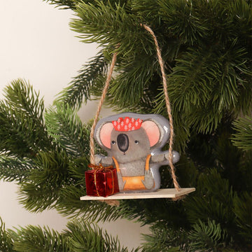 A Christmas decoration of a koala sitting on a swing holding a red present.
