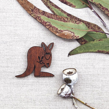 A wooden magnet in the shape of a kangaroo with a joey and gum leaves in the background.