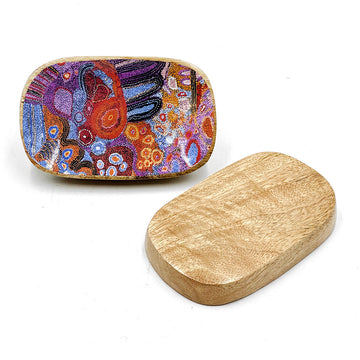 A wooden oblong trinket dish is pictured twice, one showing the patterned inside with artwork by Andrea Mimpitja Adamson and the other upside down showing the base.