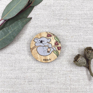 A wooden round magnet illustrated with a koala with gum leaves in the background.