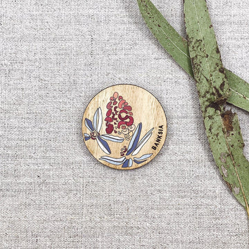 A wooden round magnet illustrated with banksia flowers with gum leaves next to the magnet.