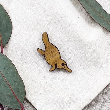 A wooden brooch shaped like a platypus with gum leaves in the background.