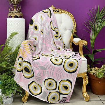 A throw blanket printed with artwork by Murdie Morris is draped over a chair with potted plants around it.