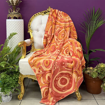 A throw blanket printed with artwork by Cedric Varcoe is draped over a chair with potted plants around it.