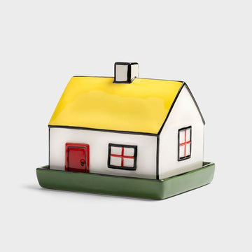 House-shaped butter dish with a yellow roof, white body, red door, and green base on a light gray background.