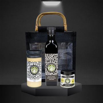 A tall jar of black garlic aioli, black garlic white balsamic dressing and a jar of black garlic with a black bag behind.