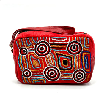A red velvet toiletry bag embroidered with artwork y Mary Napangardi Brown with a red wrist strap sitting on top.