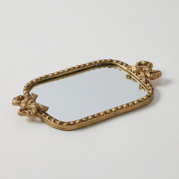 An antique gold tray with a mirror on top and embellished with bows on the ends and dots around the edge.
