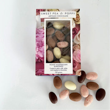 A packet filled with chocolate covered almonds with a floral border and clear window. There are loose almonds in front of the box.