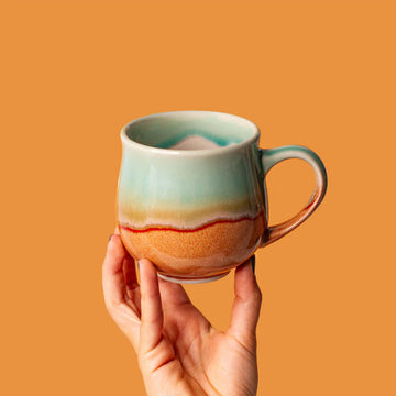 Hand holding a ceramic mug with a gradient design against an orange background.