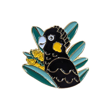 A enamel pin shaped like a black cockatoo with a wattle flower and leaves.