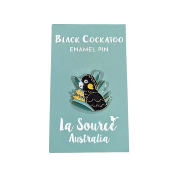 A enamel pin shaped like a black cockatoo with a wattle flower and leaves.