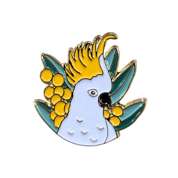 A enamel pin shaped like a white cockatoo with wattle flowers and leaves.