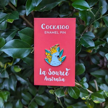 A enamel pin shaped like a white cockatoo with wattle flowers and leaves.
