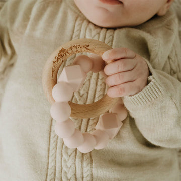 A silicone teething ring in blush pink linked to a wooden ring.