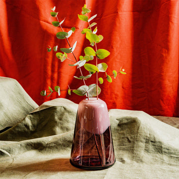 Pink vase with green leaves on a textured surface with red curtain background.