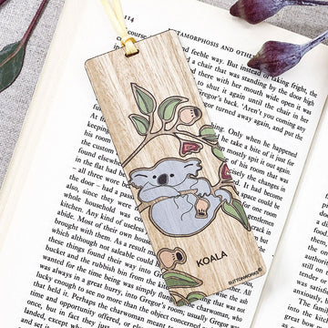 A wooden bookmark illustrated with a koala hanging off a branch inside a book.