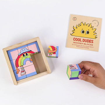 A hand is holding a wooden cube printed with different pictures next to an almost completed puzzle made from wooden cubes.