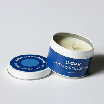 A tin with a candle inside and the lid next to the tin. The lid and tin are covered with a blue label with the text 'Lucian Mulberry and Sandalwood'.