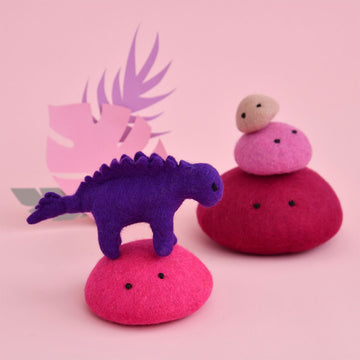 A purple stegosaurus made from felt standing on a pink felt rock. There are three pink felt rocks next to the stegosaurus.