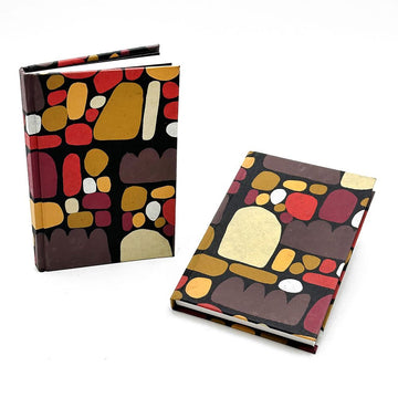 Two notebooks displayed, one standing up and one lying down. They are printed with Indigenous artwork in shades of brown, red and yellow.