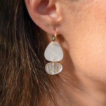 A pair of gold hoop earrings with two irregular shaped round shapes linked together in shades of white and orange.