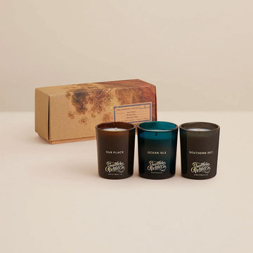 Three candles with labels in front of a branded box on a beige background.