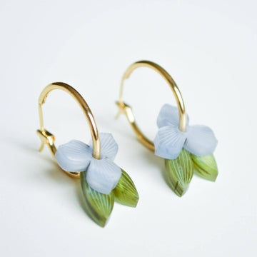 A pair of gold hoop earrings layered with a blue flowers and green leaves through the hoop.