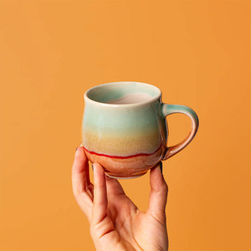 Hand holding a ceramic mug with a gradient design against an orange background.