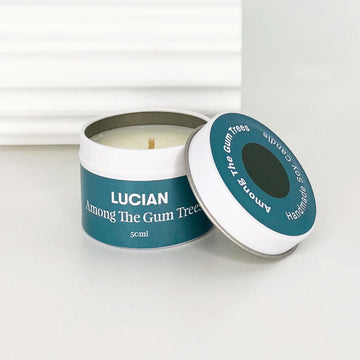 A small tin with a candle inside and the lid resting on the side. The lid and tin have a teal label with the text 'Lucian Among the Gum Trees'.