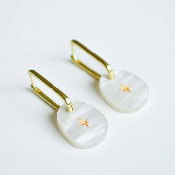 A pair of gold hoop earrings with a white coloured charm with a gold star hanging off the hoop.