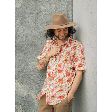 A model wears a coral pink floral short sleeve shirt with  brown pants and a brown hat while leaning against a concrete wall.