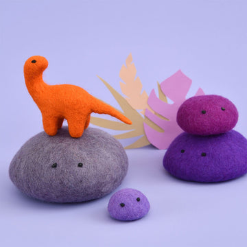 An orange brontosaurus made from felt standing on a grey felt rock. There are purple felt rocks surrounding.