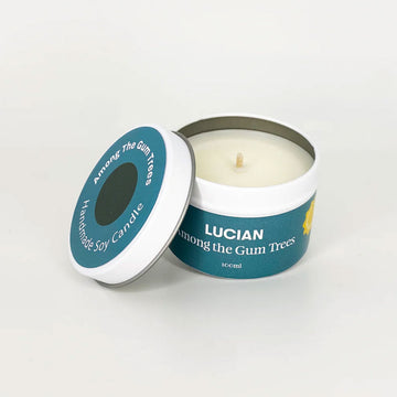 A tin with a candle inside and the lid leaning against the side. The lid and tin have a teal label with the text "Lucian Among the Gum Trees'.