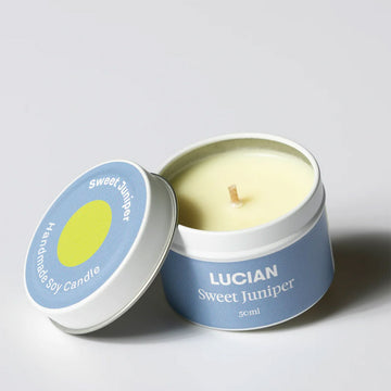 A candle in a tin with the lid resting against the side. The lid and tin have a light blue label with the text 'Lucian Sweet Juniper'.