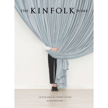 'The Kinfolk Home' book cover featuring a photo of a person standing behind a large curtain and holding it up next to the their waist. Only the person's black pants and black shoes are visible.