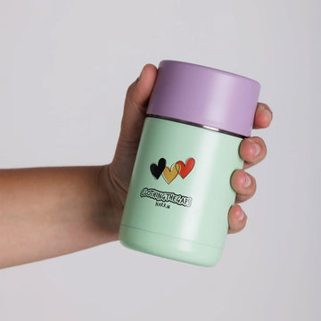 A hand is holding out a reusable green cup with a purple lid. The cup has a black, yellow and red heart on the front and the text 'Clothing the Gaps'.
