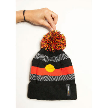 A hand is pinching the top pom pom on a beanie. The beanie has the Aboriginal flag knitted into it.
