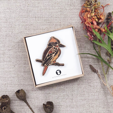 A wooden Australian kookaburra bird brooch. The Kookaburra is sitting side on, on a branch that makes up part of the brooch.  