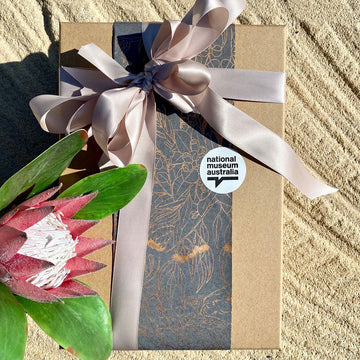 Example of gift wrapping a rectangle shape item with brown paper and pink ribbon tied into a bow over the top. A national museum sticker is on the paper.