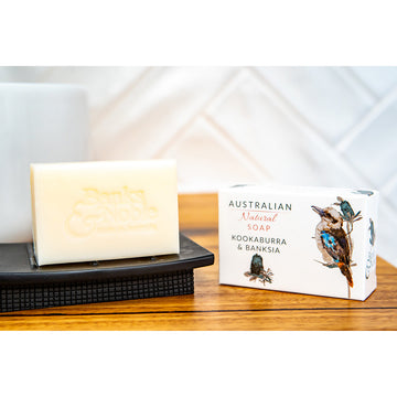 White bar of soap sitting upright on a black soap holder next to a cream box depicting an image of a kookaburra and a banksia.