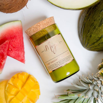 A candle made from a wine bottle with a cork lide is lying amount fruits including a mango and watermelon.