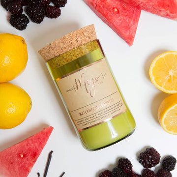 A candle made from a wine bottle with a cork lid is lying down surrounded by fruits including watermelon, berries and lemons.