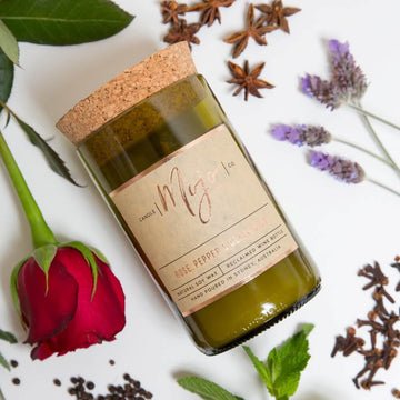 A candle made from a wine bottle with a cork lid is lying down surrounded by flowers and spices including a rose and lavender.