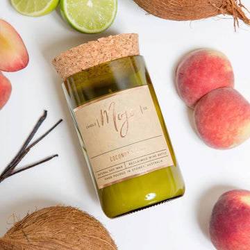 A candle made from a wine bottle with a cork lid is lying down surrounded by fruits including peaches, coconuts and limes.