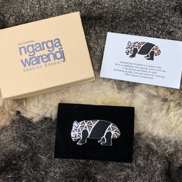 Enamel on metal brooch featuring a black, white and brown patterned Australian wombat. The wombat has black featured around the middle of the body and legs. The wombat brooch is in a standing position. Metal, enamel inlay, lapel pin by Ngarga Warendj. Features a wombat design in standing side view, open eye, and ear up. Wombat face and details in ochre brown and white.